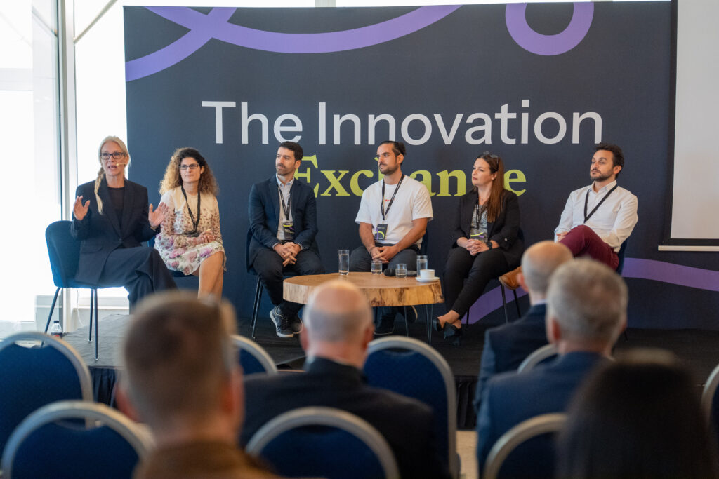 A six-person panel on stage in front of The Innovation Exchange backdrop, with the audience in the foreground.